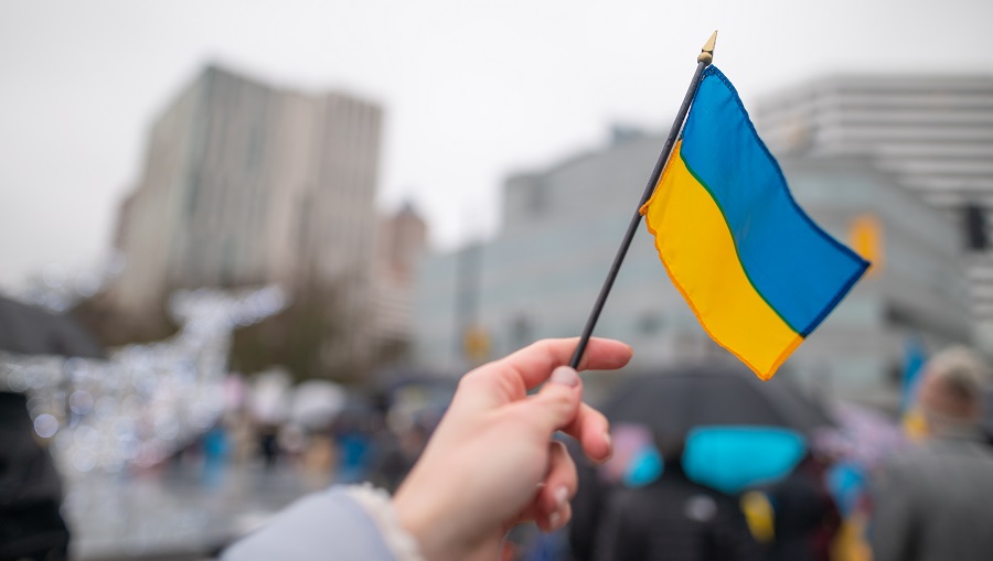 A hand holding a small Ukrainian flag in front of city buildings