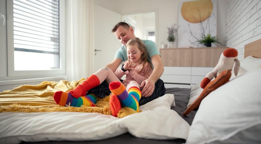 Father putting on different socks to his little daughter with Down syndrome when sitting on bed at home
